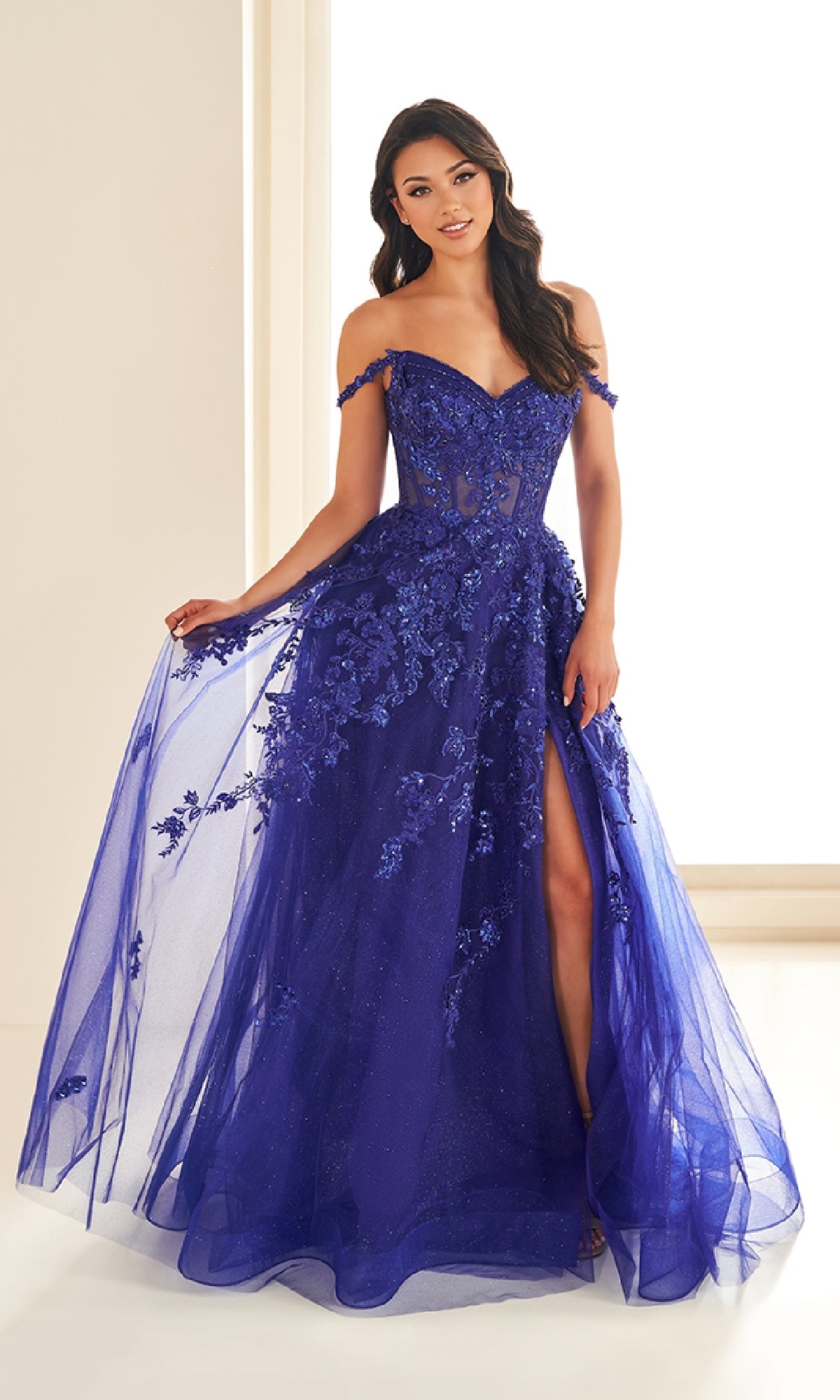 A woman stands indoors wearing the Ellie Wilde EW36204, a long royal blue prom dress with an off-the-shoulder corset bodice, floral appliqués, and a thigh-high slit.